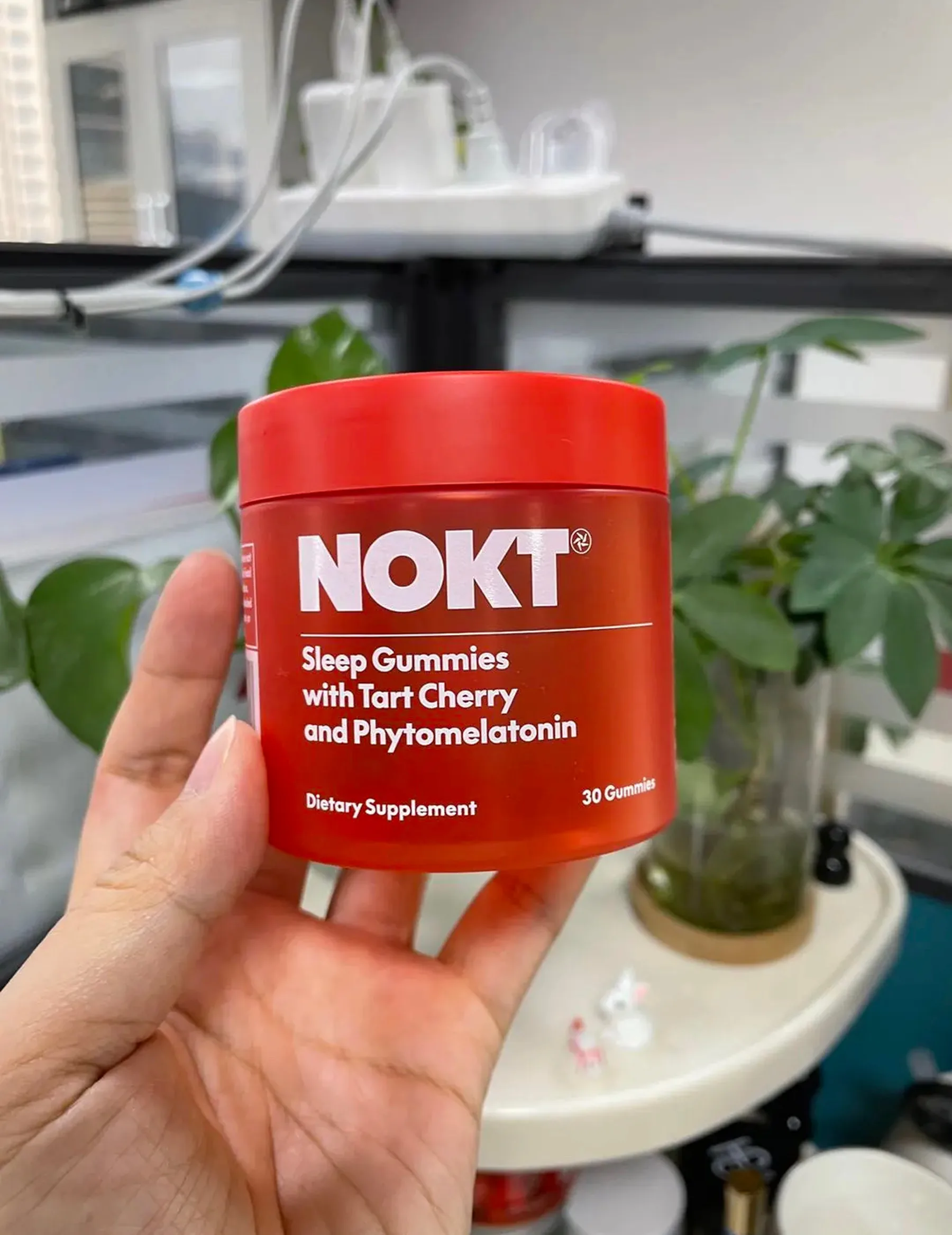 A hand holding the printed red-tinted NOKT jar with white type